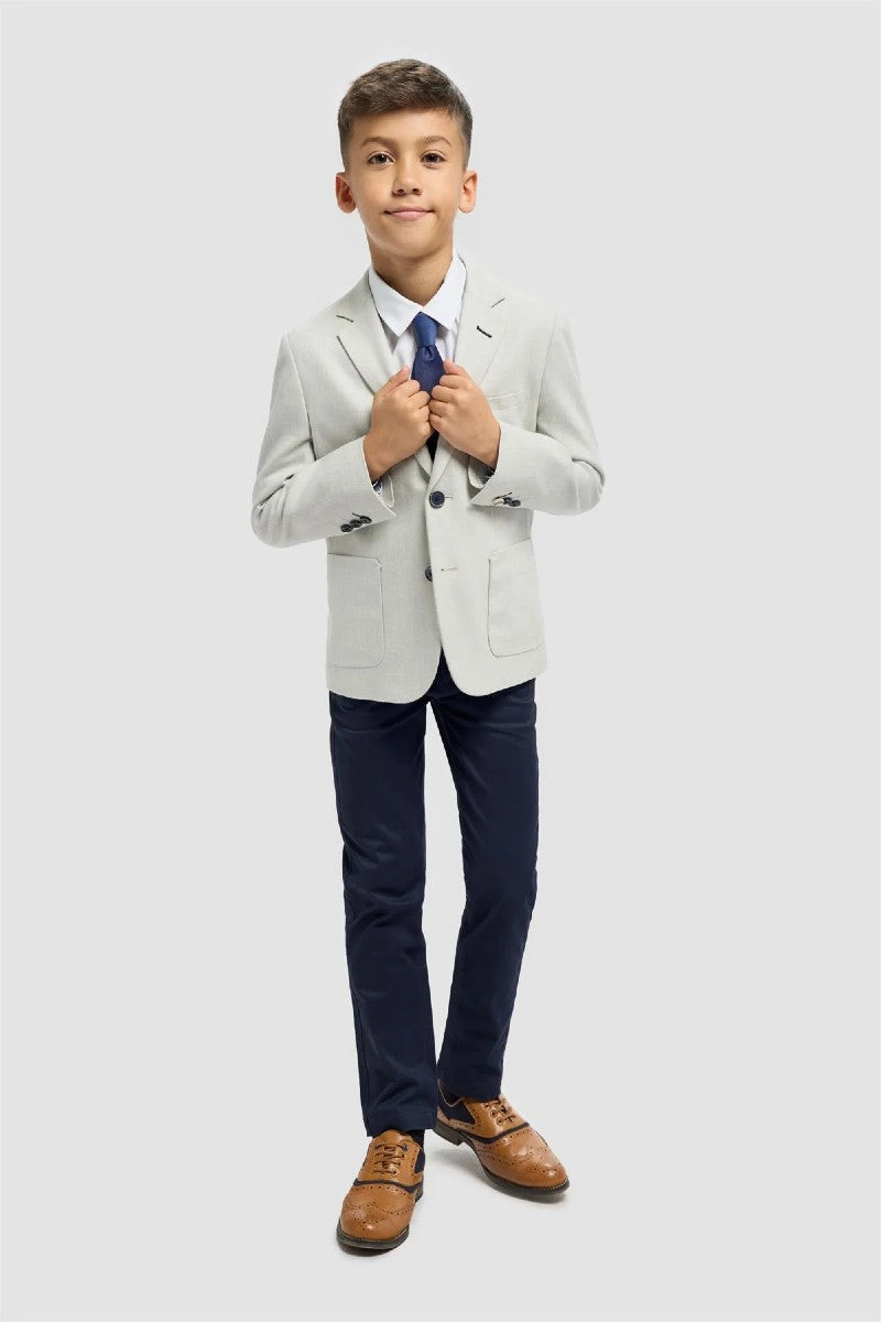 Young boy in a formal outfit with a white blazer, navy pants, and brown shoes on a light gray background