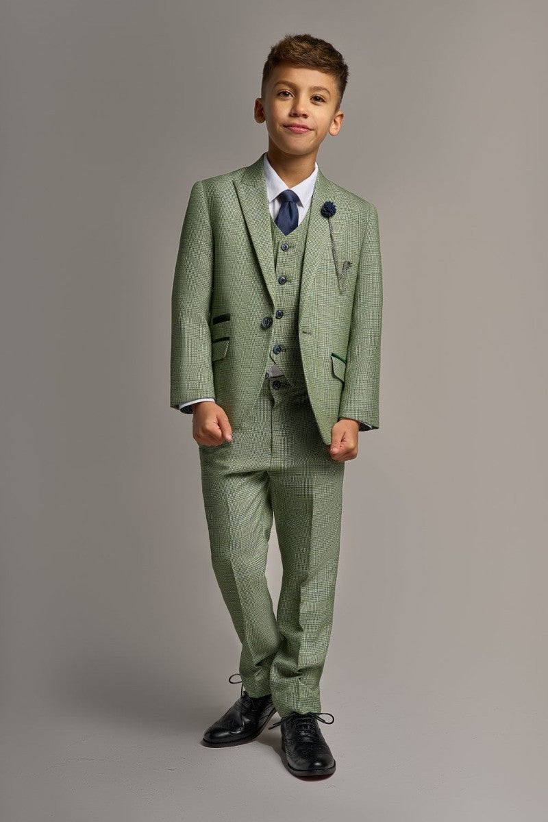 A boy wearing a sky blue three piece suit with a white shirt and a blue tie, standing against a grey background.